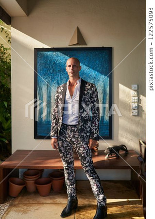 Man in bold floral suit stands confidently at villa patio. Sunlight casts shadows on wall art. Elegant gay groom attire. Trendy fashion statement. Potential wedding, fashion, lifestyle, event. 122575093