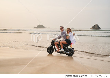 Couple rides motorbike on sandy beach at sunset. Man drives scooter with woman enjoying view. Travel fun, explore coast on budget vacation. Adventure lovers embrace leisure ocean journey together. Couple rides motorbike on sandy beach at sunset. Man drives scooter with woman enjoying view. Travel fun, explore coast on budget vacation. Adventure lovers embrace leisure ocean journey together. 122575107