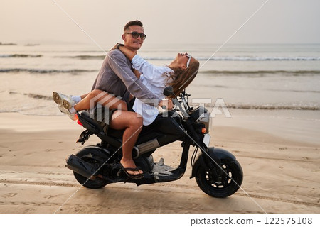 Young couple rides on motorbike by ocean beach. Man drives motorcycle while woman joyfully leans back. Lovers enjoy romantic seaside travel on scooter. Adventure at sandy coast. Young couple rides on motorbike by ocean beach. Man drives motorcycle while woman joyfully leans back. Lovers enjoy romantic seaside travel on scooter. Adventure at sandy coast. 122575108