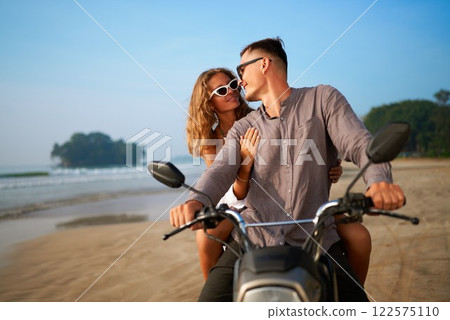 Couple rides motorcycle on beach. Man drives while woman embraces him from behind. Romantic adventure by sea. Blue sky, calm ocean, serene island backdrop. Perfect travel getaway moment. Couple rides motorcycle on beach. Man drives while woman embraces him from behind. Romantic adventure by sea. Blue sky, calm ocean, serene island backdrop. Perfect travel getaway moment. 122575110