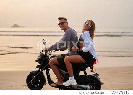 Young couple enjoys beach ride at sunset. Woman hugs man on motorbike. Romantic travel adventure on sandy shore. Freedom, love, and joy in seaside escape. Young couple enjoys beach ride at sunset. Woman hugs man on motorbike. Romantic travel adventure on sandy shore. Freedom, love, and joy in seaside escape. 122575112