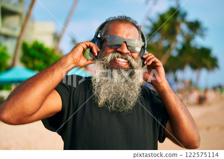 Senior bearded man listens to music on beach with sunglasses. Stylish, lively DJ enjoys ocean vibes. Modern grandpa dances with tech. Happy energetic elder at coastal party. Senior bearded man listens to music on beach with sunglasses. Stylish, lively DJ enjoys ocean vibes. Modern grandpa dances with tech. Happy energetic elder at coastal party. 122575114