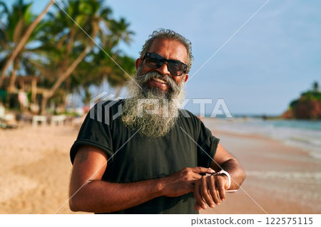 Stylish gray-bearded senior with sunglasses uses smart watch on tropical beach. Modern tech-savvy grandpa enjoys active lifestyle and sunny day. Elderly man embraces digital fitness. 122575115