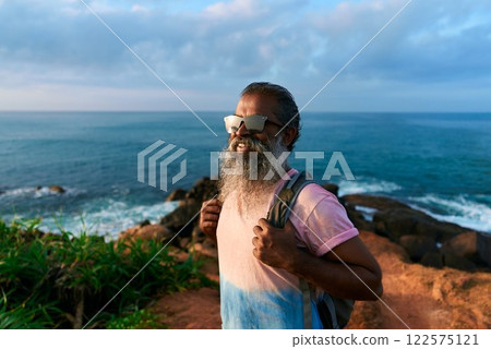 Senior black man hikes on rocky coast with backpack, sunglasses. Ocean view, blue sky. Elderly male explores nature. Active travel, outdoor adventure in retirement. Happy mature adventurer. Senior black man hikes on rocky coast with backpack, sunglasses. Ocean view, blue sky. Elderly male explores nature. Active travel, outdoor adventure in retirement. Happy mature adventurer. 122575121