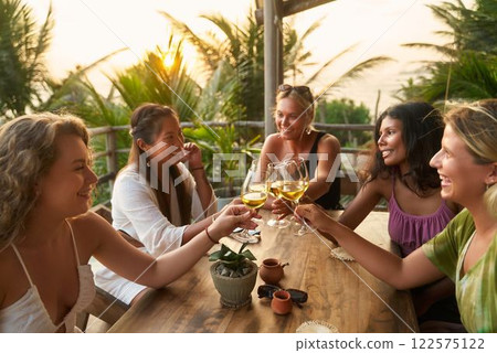 Diverse women toast with glasses at tropical restaurant. Friends share drinks, enjoy bachelorette party, smile. Relaxation, fun atmosphere. Wine tasting at sunset by ocean view. 122575122