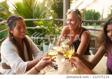 Group of diverse women toasting with wine in restaurant. Friends gather, laugh, celebrate social event. Multicultural female meeting. Stylish casual dresses. Cheers on terrace with nature view. 122575124