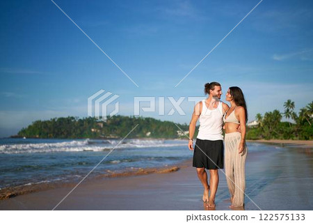 Romantic couple walks on sandy beach at sunset. Barefoot man, woman in casual summer attire hold hands, enjoying seaside honeymoon stroll. Tranquil ocean scenery, palm trees, vacation lovers leisure. 122575133