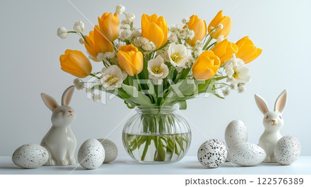 Yellow and white tulips in a glass vase, paired with speckled eggs and ceramic rabbits on a table. 122576389