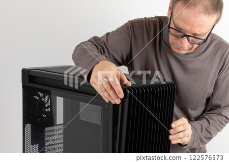 Man removing  15.5 mm front panel grille with 45% porosity with dust filter from  PC case, empty big tower, chassis, on side is tempered-glass. 122576573