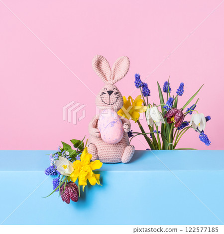 Knitted toy pink bunny with Easter egg and bouquet of spring flowers on blue table and purple background. Concept of spring, Easter, Mother's Day 122577185