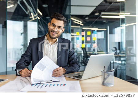 Portrait of successful financier, man signs documents reports contracts. Businessman at workplace inside office doing paper work, filling out tax and statistical forms, looking at camera smiling. Portrait of successful financier, man signs documents reports contracts. Businessman at workplace inside office doing paper work, filling out tax and statistical forms, looking at camera smiling. 122579857