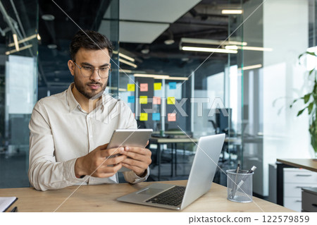 Serious thinking businessman working with tablet computer inside office. Businessman in shirt working at workplace, using laptop, sitting at desk, using financial app. Serious thinking businessman working with tablet computer inside office. Businessman in shirt working at workplace, using laptop, sitting at desk, using financial app. 122579859