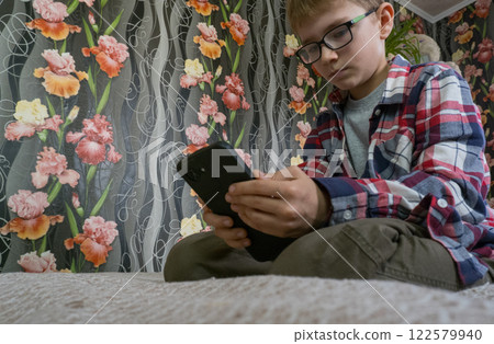 A boy on the sofa with a mobile phone in his hands. The boy is addicted to gadgets and uses a mobile phone at home. Internet obsession concept. A boy on the sofa with a mobile phone in his hands. The boy is addicted to gadgets and uses a mobile phone at home. Internet obsession concept. 122579940