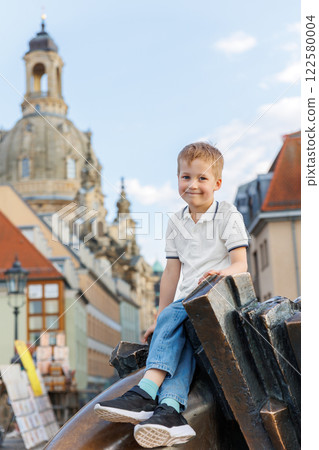 Smiling little kid boy sits on bronze sculpture  against Frauenkirche grand baroque dome  ornate spire gracefully rising behind him in Dresden Germany.  Family European city travel destination 122580004