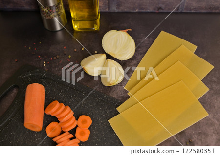 Carrot on the black kitchen cutting board. Lasagne pasta sheets, onion, oil, pepper. Black background. Homemade lasagna cooking preparation Carrot on the black kitchen cutting board. Lasagne pasta sheets, onion, oil, pepper. Black background. Homemade lasagna cooking preparation 122580351