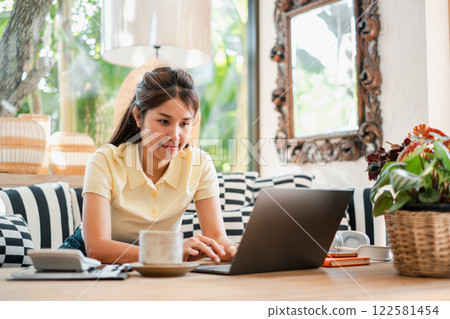 A young woman focused on her laptop in a cozy, well-decorated living room with natural light and modern furnishings. 122581454