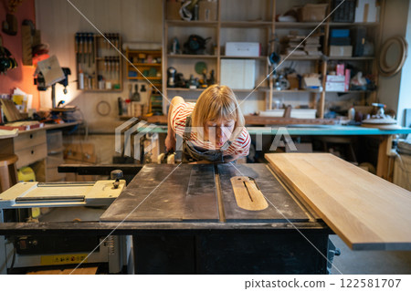 Female handywoman blowing dust off table surface before starting circular saw work in workshop Female handywoman blowing dust off table surface before starting circular saw work in workshop 122581707