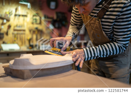 Woodworker carefully smooths stool base in carpentry studio. Surface preparation, manual sanding 122581744