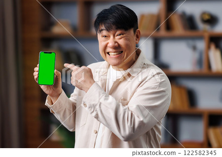 happy asian man, showing device, Online offer, empty free space, man with short hair is excitedly pointing at a smartphone displaying a blank screen. He stands in a welcoming indoor space filled with 122582345