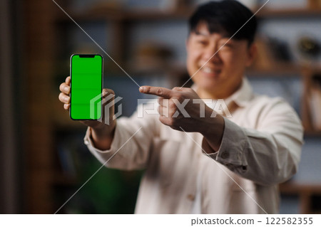 Individual is happily showcasing a smartphone with a vibrant green display in a warm, inviting room filled with personal items. The gesture highlights the device's screen. Individual is happily showcasing a smartphone with a vibrant green display in a warm, inviting room filled with personal items. The gesture highlights the device's screen. 122582355