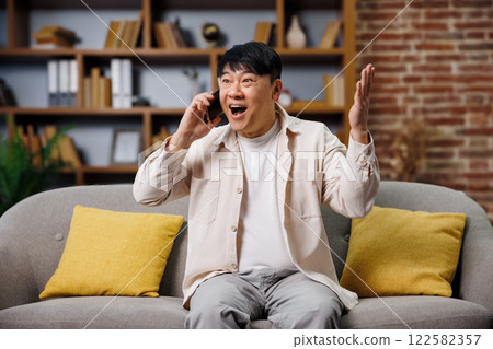hear good news, talking phone, Excited man sits on a gray couch in a stylish living room, animatedly talking on the phone with a big smile, expressing excitement and joy in a relaxed atmosphere. hear good news, talking phone, Excited man sits on a gray couch in a stylish living room, animatedly talking on the phone with a big smile, expressing excitement and joy in a relaxed atmosphere. 122582357