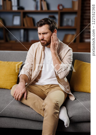 severe toothache, Upset man, holds his cheek, man appears to be in discomfort while sitting on a couch in a well-decorated living room. He holds his ear with a concerned expression, suggesting he may 122582368