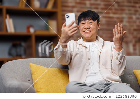 video call conversation, telephone conversation, smiling asian man is smiling while engaging in a video call from a comfortable couch, surrounded by modern home decor. It is a sunny afternoon video call conversation, telephone conversation, smiling asian man is smiling while engaging in a video call from a comfortable couch, surrounded by modern home decor. It is a sunny afternoon 122582396