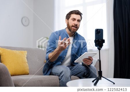 conference work meeting, remote job interview, online broadcast, lesson, man is actively participating in a virtual meeting on his tablet, sitting comfortably on a couch in a bright living room filled 122582413