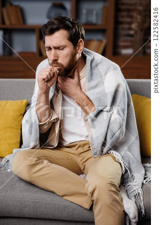 man coughs, feeling unwell, cold throat, man sits on a couch, wrapped in a blanket, displaying signs of illness as he coughs. The cozy living room features warm lighting and yellow pillows, creating a 122582416