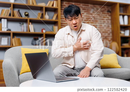 working man, having heart attack, chest heartache, man displays signs of chest discomfort while seated on a cozy couch, glancing at a laptop in a stylish living room filled with bookshelves and working man, having heart attack, chest heartache, man displays signs of chest discomfort while seated on a cozy couch, glancing at a laptop in a stylish living room filled with bookshelves and 122582420