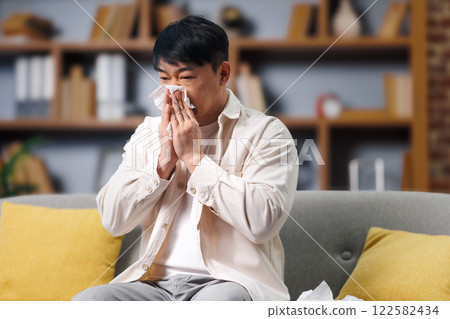 ill sick man, feeling unwell, having headache, man sits on a comfortable couch with yellow cushions, wiping his nose with a tissue. He appears to be struggling with a cold or allergies amid a cozy ill sick man, feeling unwell, having headache, man sits on a comfortable couch with yellow cushions, wiping his nose with a tissue. He appears to be struggling with a cold or allergies amid a cozy 122582434