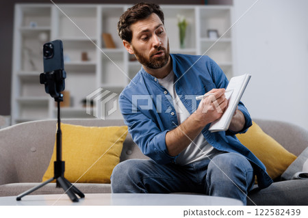 conference work meeting, remote job interview, online broadcast, lesson, man sits on a couch in a stylish living room, writing in a notebook while using a smartphone for reference. He appears focused conference work meeting, remote job interview, online broadcast, lesson, man sits on a couch in a stylish living room, writing in a notebook while using a smartphone for reference. He appears focused 122582439