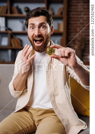 excited man, shows cryptocurrency, gold bitcoin coin, man with a beard and cheerful expression holds up a bitcoin in a well-decorated living room. His excitement reflects the growing interest in 122582468