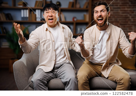fans watching game, celebrating victory, Excited friends watching TV, Two friends express delight after a sports victory, sitting on a comfortable couch in a cozy living room filled with books and 122582478