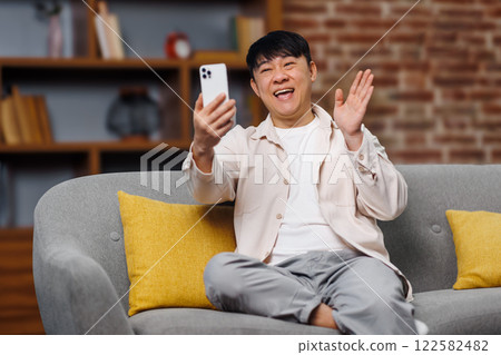 video call conversation, telephone conversation, smiling asian man, cheerful man is engaged in a video call from his cozy living room. He smiles and waves while sitting on a soft couch adorned with video call conversation, telephone conversation, smiling asian man, cheerful man is engaged in a video call from his cozy living room. He smiles and waves while sitting on a soft couch adorned with 122582482