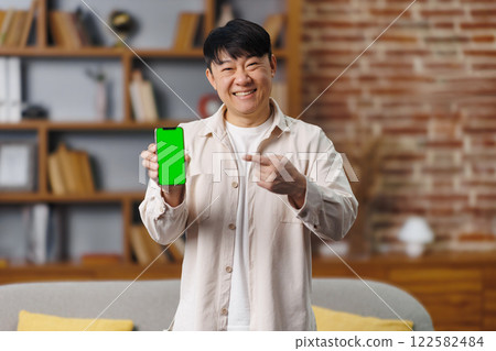 happy asian man, showing device, Online offer, empty free space, cheerful man points to a smartphone displaying a vibrant green screen while standing in a stylish living room with bookshelves and warm happy asian man, showing device, Online offer, empty free space, cheerful man points to a smartphone displaying a vibrant green screen while standing in a stylish living room with bookshelves and warm 122582484