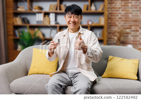 glass water, smiling happy man, thumb up, cheerful man sits comfortably on a gray sofa with yellow cushions, holding a glass of drink and showing a thumbs up, enjoying the cozy atmosphere of his 122582492