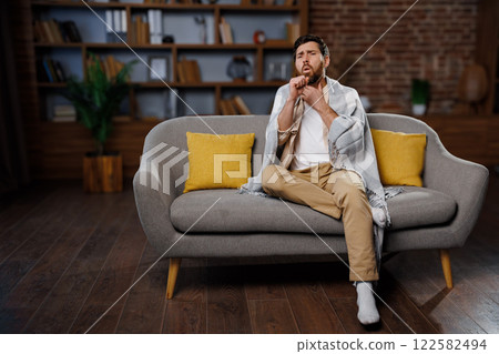 man coughs, feeling unwell, cold throat, man sits comfortably on a gray sofa, wrapped in a soft blanket, with bright yellow cushions nearby. He appears deep in thought, reflecting on personal 122582494