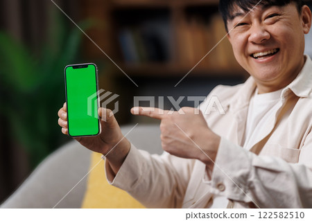 Amazing Useful App, Great Mobile Offer, Mobile Advertisement, Excited man, cheerful man points at a smartphone with a green screen, sitting comfortably in his living room decorated with plants 122582510