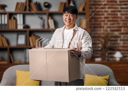 opening delivery box, excited man, Online shopping concept, man is standing in a welcoming living room, cheerfully holding a large cardboard box. The room features bookshelves filled with books opening delivery box, excited man, Online shopping concept, man is standing in a welcoming living room, cheerfully holding a large cardboard box. The room features bookshelves filled with books 122582513