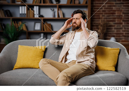temples pain,having headache, massaging temples, man in casual attire is seated on a gray sofa, with yellow cushions, appearing to struggle with stress or discomfort in a stylishly decorated living 122582519