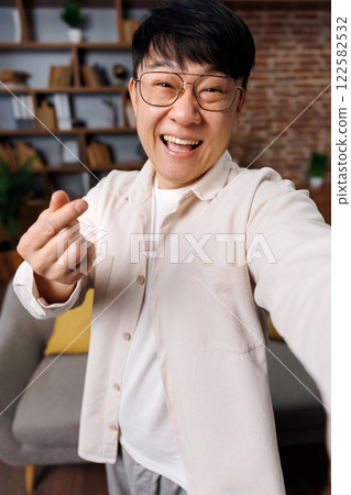shows sign love, takes selfie, video call, joyful person stands in a stylish living room, smiling broadly while forming a heart shape with their fingers, creating a friendly and inviting vibe in the shows sign love, takes selfie, video call, joyful person stands in a stylish living room, smiling broadly while forming a heart shape with their fingers, creating a friendly and inviting vibe in the 122582532
