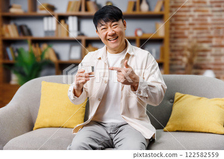 A cheerful senior man seated on a soft sofa holds a glass of water, smiling broadly while pointing to it. The warm interior features bookshelves and bright cushions, creating a welcoming atmosphere. 122582559
