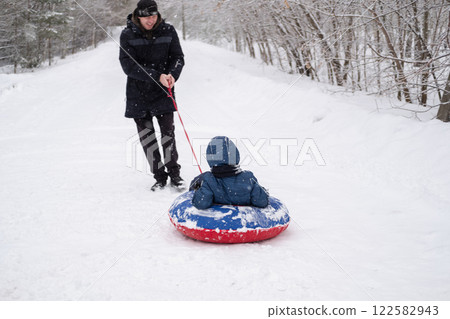 dad in winter jacket rides child on snow tubing around himself, against the background of snowfall and forest in winter. Dangerous and fun games of dad and son in winter dad in winter jacket rides child on snow tubing around himself, against the background of snowfall and forest in winter. Dangerous and fun games of dad and son in winter 122582943