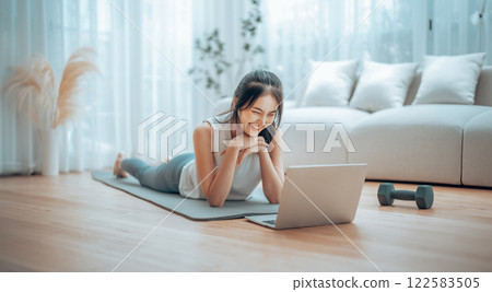 Young Asian woman lying on the floor on yoga mat in front of laptop and training at home 122583505