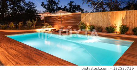 Illuminated backyard swimming pool with wooden deck and lounge chairs. Serene outdoor space surrounded by greenery and soft lighting 122583739