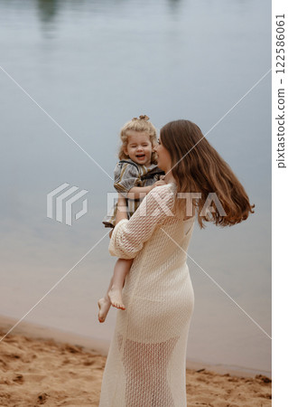 Joyful Caucasian mother in beige dress lifts smiling daughter into air on lake shore. 122586061