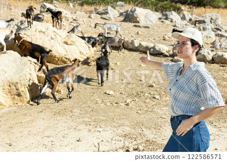 Female shepherd manages herd of goats on rough terrain on summer day 122586571
