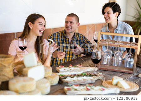 One man and two women enjoying degustation of cheese while holding glasses with red wine on rustic wooden table in restaurant 122586912