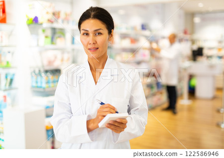 Positive female pharmacist makes notes on a notebook in the sales area of pharmacy Positive female pharmacist makes notes on a notebook in the sales area of pharmacy 122586946